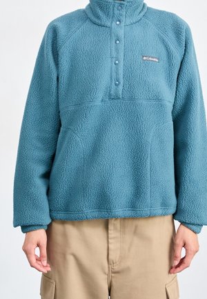 Blå Columbia fleece-pullover med trykknapper og høj krave, båret med beige cargobukser, forfra set fra skuldre til hofter.