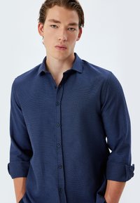 Marineblaue Button-down-Hem aus strukturiertem Stoff mit Kragen und umgeschlagenen Ärmeln. Verfügt über sechs Knöpfe und eine normale Passform.
