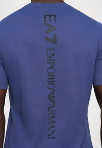 Zils t-krekls ar vertikālu melnu "EA Emporio Armani" logo un emblemu izdrukātu pa vidu aizmugurē.