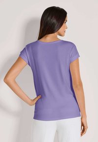 Lila paarse t-shirt met korte mouwen en een ronde halslijn, gemaakt van een gladde stof, gedragen met witte broek en toont een casual pasvorm.