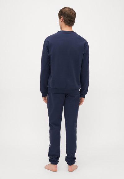 Mann som står barbeint med ryggen til, iført matchende marineblå hettegenser og joggebukser mot en enkel hvit bakgrunn.