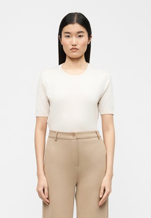 Femme aux cheveux noirs raides portant un haut blanc ajusté à manches courtes, rentré dans un pantalon beige taille haute, sur un fond uni.