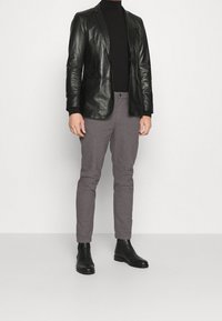 Man i svart läderblazer, svart turtleneck, grå texturerade byxor och svarta ankelboots, står mot en enkel ljus bakgrund.