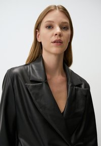 Zwarte leren blazer met een diepe V-hals en opvallende revers. De stof heeft een gladde textuur en een getailleerd ontwerp.