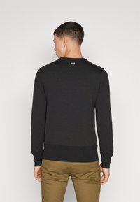 Schwarzer, gerippter Pullover mit Rundhalsausschnitt, elastischen Bündchen und Saum. Besitzt ein kleines Logotäfelchen am Nacken. Model trägt khakifarbene Hose.