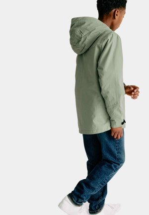 Giacca con cappuccio verde chiaro, dalla texture liscia, abbinata a jeans blu scuro e sneakers bianche, vista da dietro.