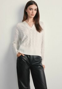 Biały sweter w splot warkoczowy z dekoltem w kształcie V i prążkowanym wykończeniem, zestawiony z czarnymi skórzanymi spodniami. Gładka tekstura, luźny krój i długie rękawy.