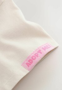 Flødefarvet stof med en blød tekstur; har en pink syet etiket med teksten "ADOPT ME!" på kanten.