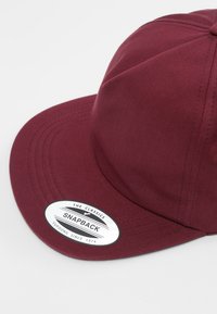 Burgundi snapback sapka lapos peremmel, texturált anyagból készült, fehér kör alakú folttal, amelyen a "The Classics SNAPBACK" felirat található.