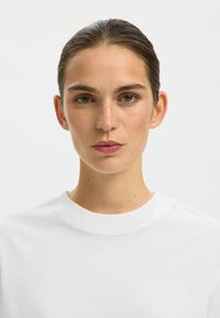 Valge ümmarguse kaelusega T-särk lühikeste varrukatega, sile puuvillane tekstuur ja minimalistlik disain. Modellil on taha kammitud juuksed ja naturaalne meik.