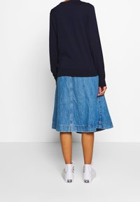 Personne portant un haut bleu marine à manches longues, une jupe en jean bleu mi-longue et des baskets montantes blanches, debout devant un fond blanc.