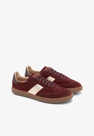 Piros velúr edzőcipők fényes arany csíkkal, barna bőr részletekkel és texturált gumi talppal. Fűzős kialakítás párnázott bokarészekkel.