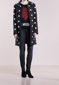 Manteau noir à pois blancs, fermeture à boutons, et grand col ; porté sur un pull noir avec un cœur rouge pailleté. Jean gris ajusté et bottines noires.