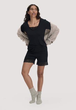 Donna che indossa pantaloncini e top neri con bordatura in pizzo, calzini grigi e un cardigan beige drappeggiato sulle spalle, in piedi su una gamba.