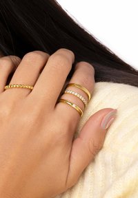 Anillos de oro en varios estilos: uno delgado con cuentas, dos bandas lisas, uno con piedras transparentes. Mano descansando sobre una tela de punto de color claro.