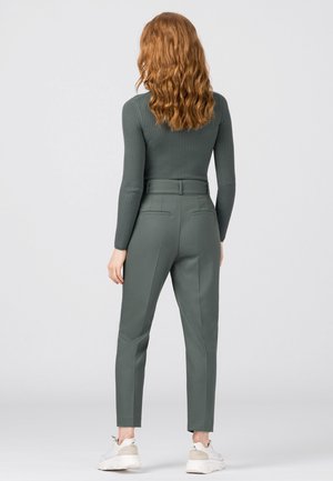 Donna con capelli rossi mossi vista di spalle che indossa una maglia verde a coste a maniche lunghe, pantaloni verdi a vita alta e scarpe da ginnastica bianche su sfondo neutro.