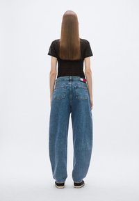 Mėlynos džinsinės kelnės su aukšta apačia ir laisvu kirpimu, turinčios dvi galines kišenes. Juosmens juostoje yra raudonos, baltos ir mėlynos spalvos etikėtė, o audinys yra lygus.