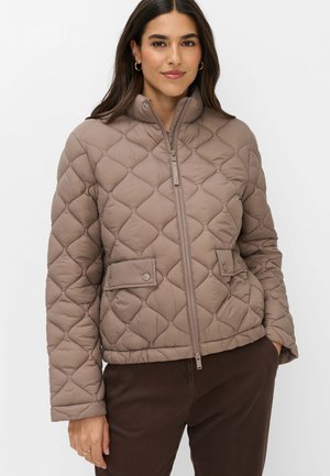 Femme portant une veste matelassée marron avec une fermeture éclair à l'avant et deux poches à rabat, associée à un pantalon marron foncé, debout sur un fond blanc.