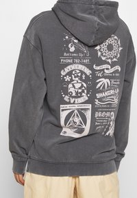 Grå huvtröja med detaljerade grafiska tryck på ryggen i vitt, inklusive text och geometriska mönster. Tillverkad i bomull, avslappnad passform.