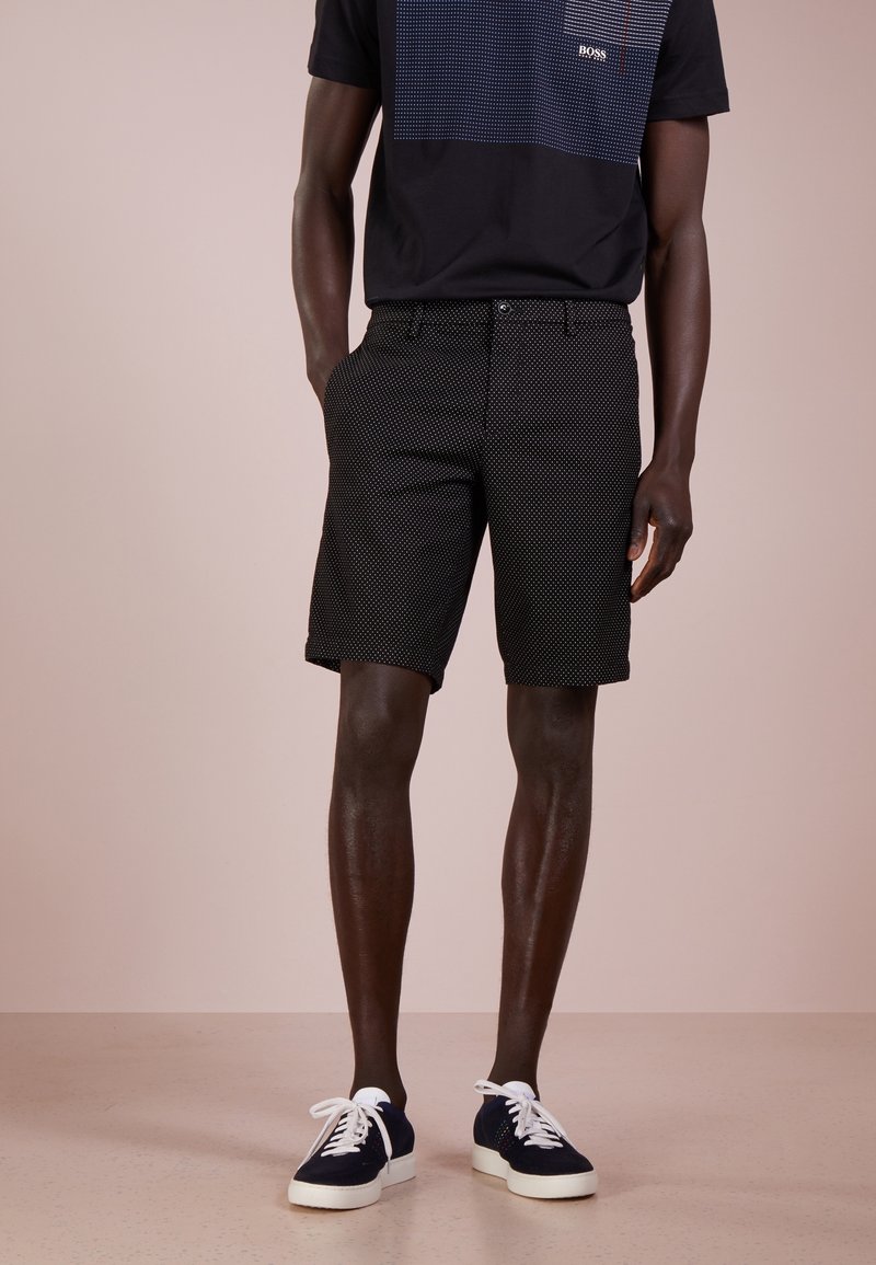 Homme portant un short noir à motif, des baskets noires et bleues avec des lacets blancs, et un t-shirt noir avec un carré à motif et un logo.