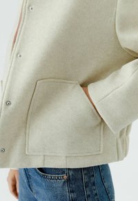 Beige Wolljacke mit Metall-Druckknöpfen, versehen mit einer großen Fronttasche und gekrempelten Ärmeln, getragen zu blauen Jeans aus Denim.