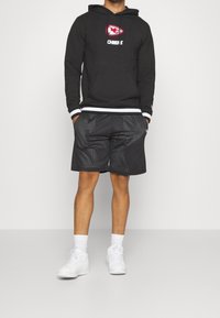Haut à capuche noir avec le logo des Kansas City Chiefs, associé à un short noir texturé avec un design en mesh, et des baskets blanches.