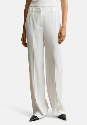 Femme portant un pantalon blanc taille haute à jambes larges avec des plis sur le devant et des chaussures noires pointues, debout devant un fond uni.