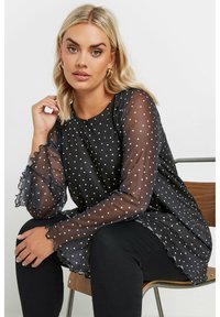 Blusa nera a pois con maniche lunghe trasparenti e bordi ondulati, abbinata a pantaloni neri. Il tessuto è leggero e fluido.
