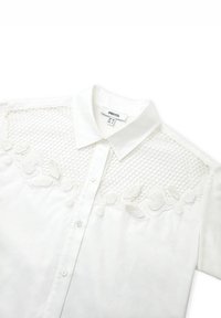 Weiße Bluse mit Kragen, Knopfverschluss und aufwendigen Spitzenverzierungen mit floralem Muster im oberen Bereich. Glatte Textur.