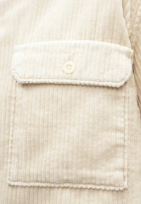 Krém színű pamut kordbársony anyag, amelyen egy gombbal záródó mellzseb és finom, függőleges bordázott textúra található.