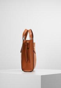 Sac fourre-tout en cuir marron avec double poignée et tirette de fermeture éclair, debout verticalement sur un piédestal blanc contre un fond uni.