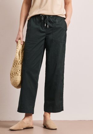 Person iført sorte vidbenede bukser med snøre, beige top, holder en vævet beige tote-taske og bærer beige slip-on sko.