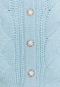 Vaaleansininen neuleliivi, jossa on letkusuunnittelu ja kolme pyöreää, kimaltavaa kristallinappia edessä.