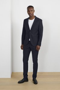 Costume marine avec un blazer à un bouton et un pantalon assorti, associé à un t-shirt blanc et des chaussures noires. Tissu lisse, coupe ajustée.