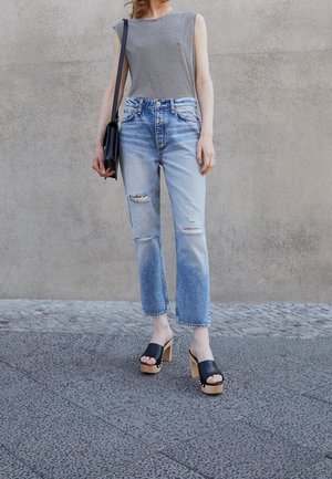 Personne portant un haut sans manches rayé noir et blanc, un jean déchiré bleu clair, des sandales noires à plateforme, et portant un sac à bandoulière noir.