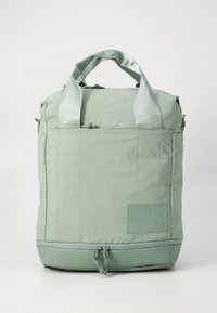 Zaļganzaļa mugursoma ar divām rokturēm, rāvējslēdzēja aizdari un izteiktu logotipa plāksteri. Izgatavota no viegla, teksturēta auduma ar auklas akcentiem.