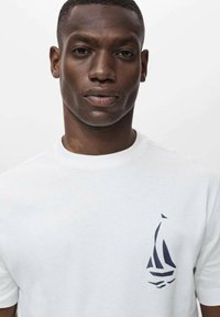 White cotton t-shirt featuring a navy blue sailboat graphic on the left chest. Short sleeves and a standard crew neckline. Smooth texture.