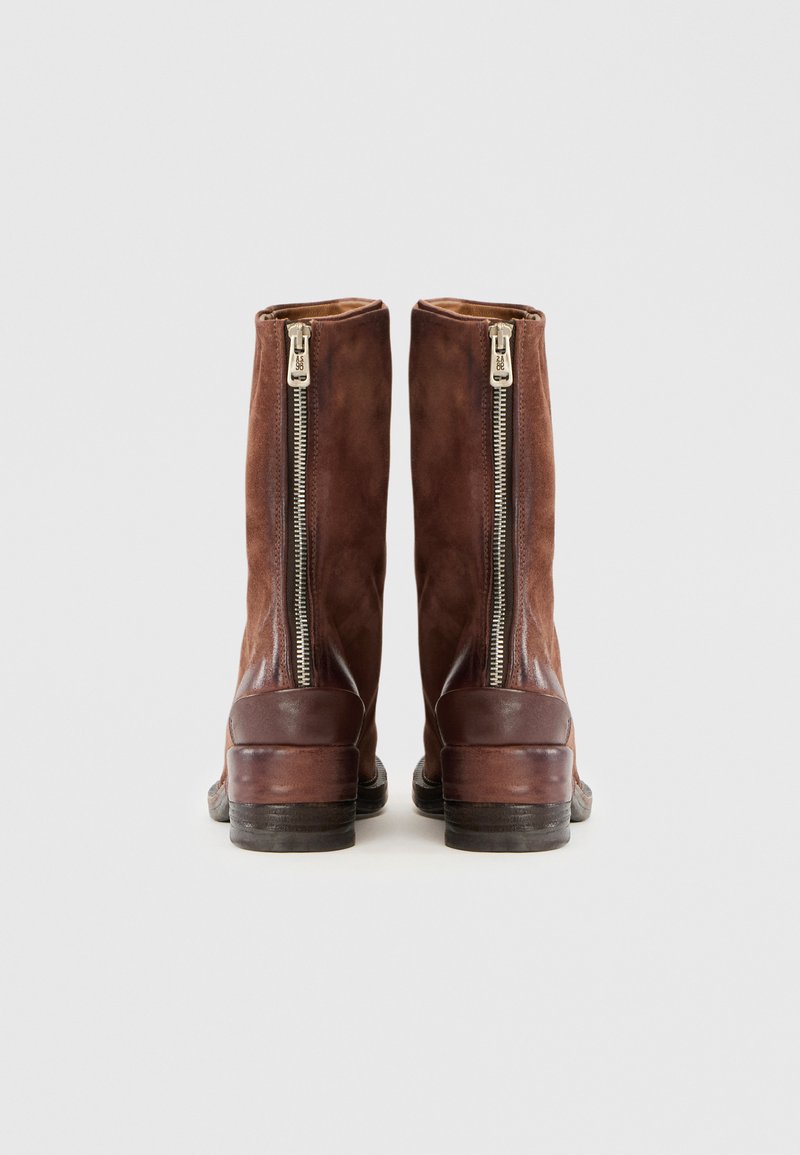 Une paire de bottes en suède marron mi-mollet avec des zippers argentés à l'arrière et des talons en bois empilés sur un fond blanc uni.
