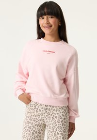 Joven mujer sonriente con cabello largo y oscuro lleva una sudadera rosa clara con el texto "FIELD OF DREAMS" y pantalones crema con estampado de leopardo, con una mano en el bolsillo.