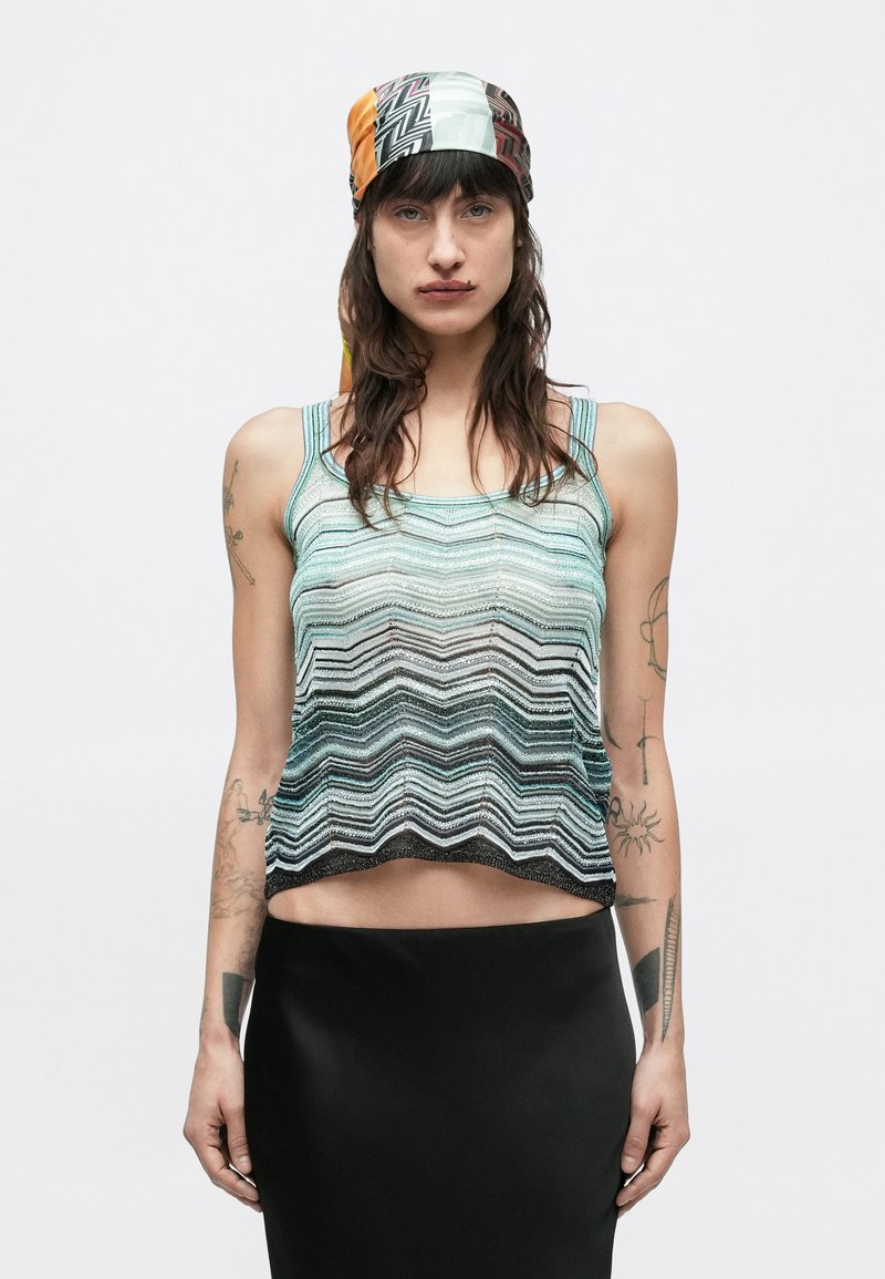 Γυναίκα με μακριά κυματιστά μαλλιά και τατουάζ φ�οράει κεφαλόδεσμο με σχέδια, κοντομάνικη μπλούζα με ζιγκ ζαγκ ρίγες και μαύρη φούστα σε απλό φόντο.