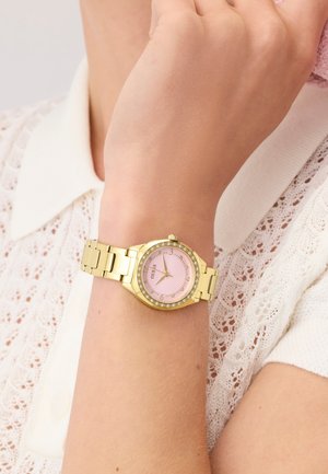 Montre-bracelet en ton or avec un cadran rose et une lunette en cristal portée sur le poignet gauche par-dessus une chemise blanche à col en dentelle.