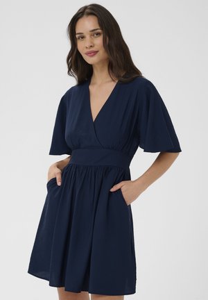 Žena s dlhými tmavými vlasmi v tmavomodrých šatách s véčkovým výstrihom, širokými rukávmi a vreckami, stojaca na jednoduchom pozadí.