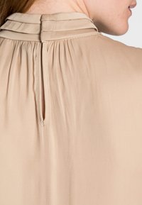 Beige bluse med høj krave, subtile plisseringer og en nøglehulåbning bagpå. Glat stof med en let tekstur.