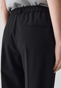 Zwarte broek met een gladde textuur, voorzien van een elastische tailleband en een achterzak. Het ontwerp is op maat gemaakt en benadrukt een moderne pasvorm.