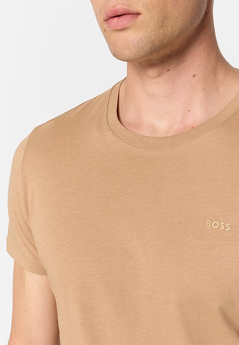 Nahaufnahme einer Person, die ein beiges T-Shirt mit Rundhalsausschnitt trägt, auf dessen Brust das gestickte "BOSS"-Logo zu sehen ist.