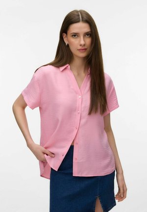 Ung kvinde med langt, glat brunt hår iført en løs, lyserød kortærmet skjorte med knapper og et blåt denimskørt med slids foran.