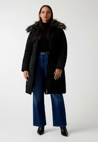 Schwarzer Steppmantel mit kapuzenbesatz aus Fell, langen Ärmeln, Reißverschluss und Seitentaschen. Kombiniert mit blauen Schlagjeans und schwarzen Stiefeletten.