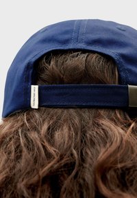 Marineblauwe pet met een verstelbare band aan de achterkant, voorzien van een klein label met de tekst "THINKING MU" nabij de band. Gelaagde stof met zichtbare stiksels.