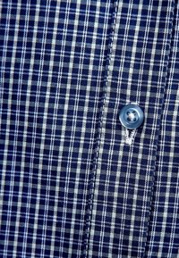 Tissu à carreaux bleu et blanc avec un gros plan sur un bouton bleu cousu et des coutures détaillées le long de la patte de boutonnage.