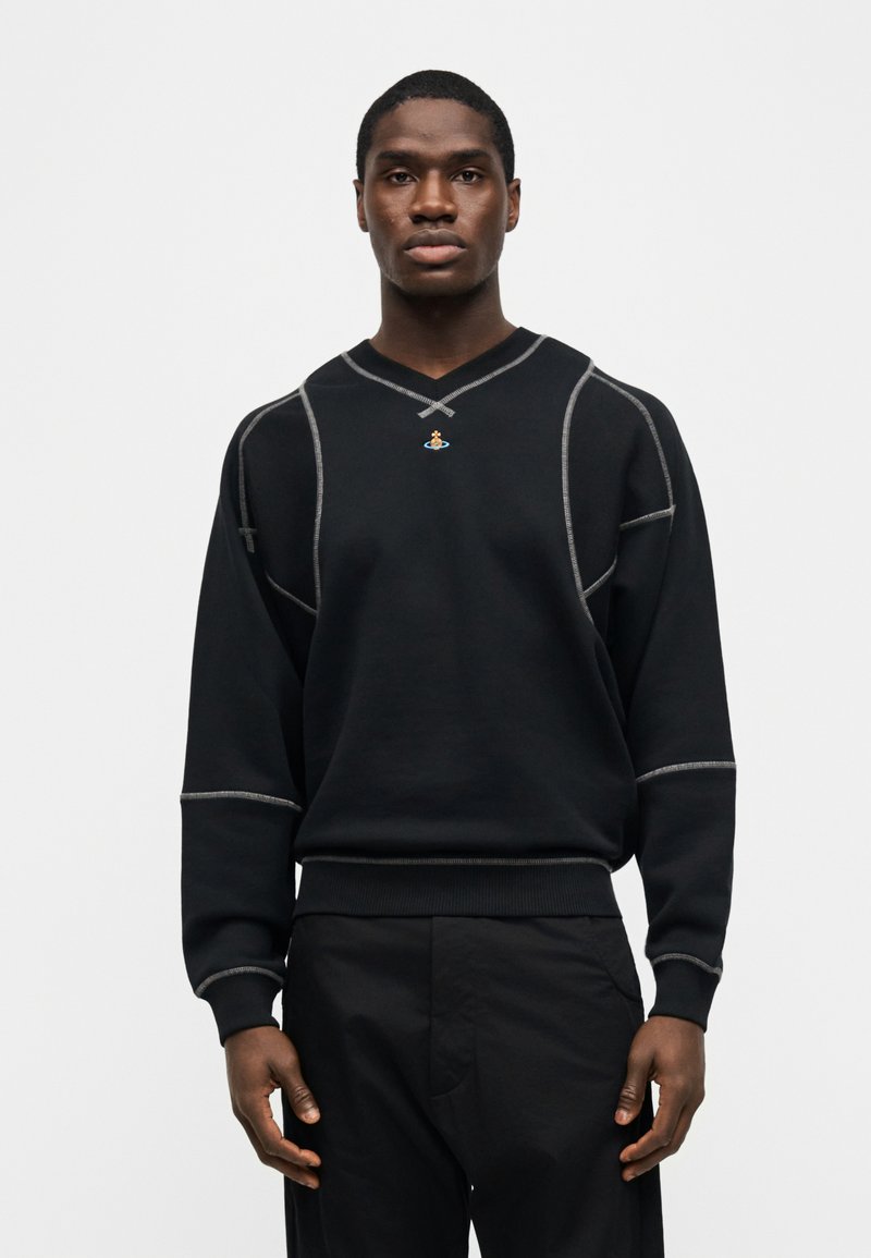 Uomo che indossa un maglione nero con dettagli cuciti bianchi e un piccolo emblema colorato sul petto, abbinato a pantaloni neri, in piedi davanti a uno sfondo bianco.